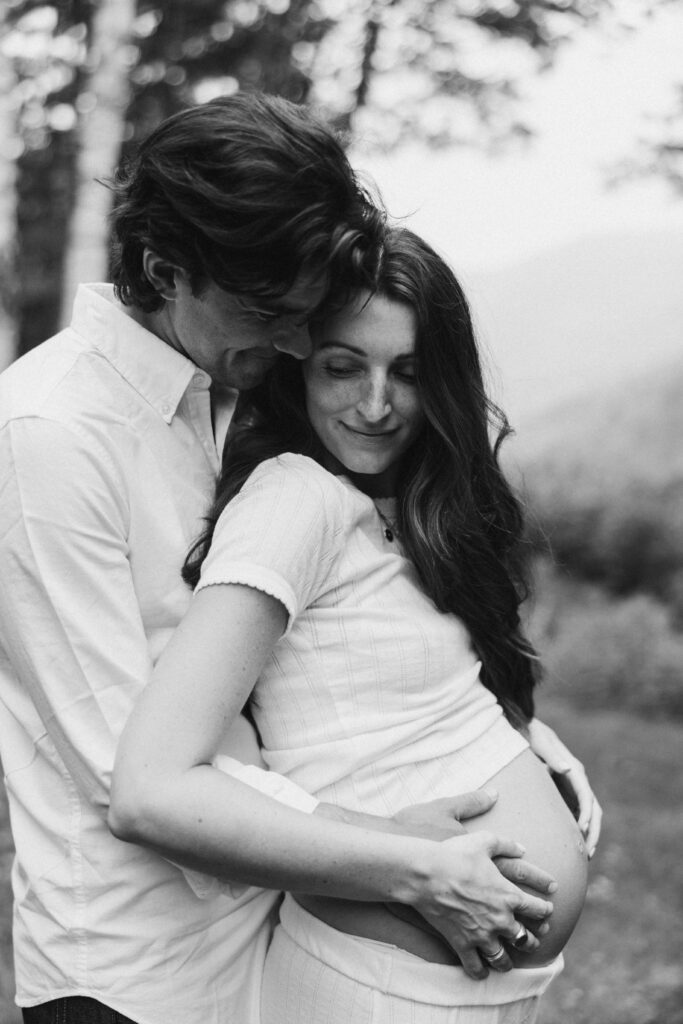 A black and white photo of a pregnant woman and her partner standing closely together in an outdoor setting. The man lovingly embraces her from behind, his hands gently resting on her belly, while they both share a tender, serene smile. The natural background is softly blurred, emphasizing their intimate connection.