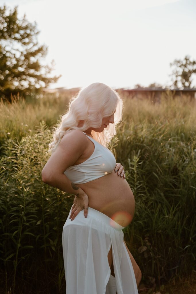 A pregnant woman stands in a sunlit field, gently cradling her belly with one hand while supporting her back with the other. She wears a white crop top and flowing skirt, her long blonde hair cascading in soft waves. The golden hour light creates a warm and ethereal atmosphere, capturing the serenity and beauty of this maternity moment.