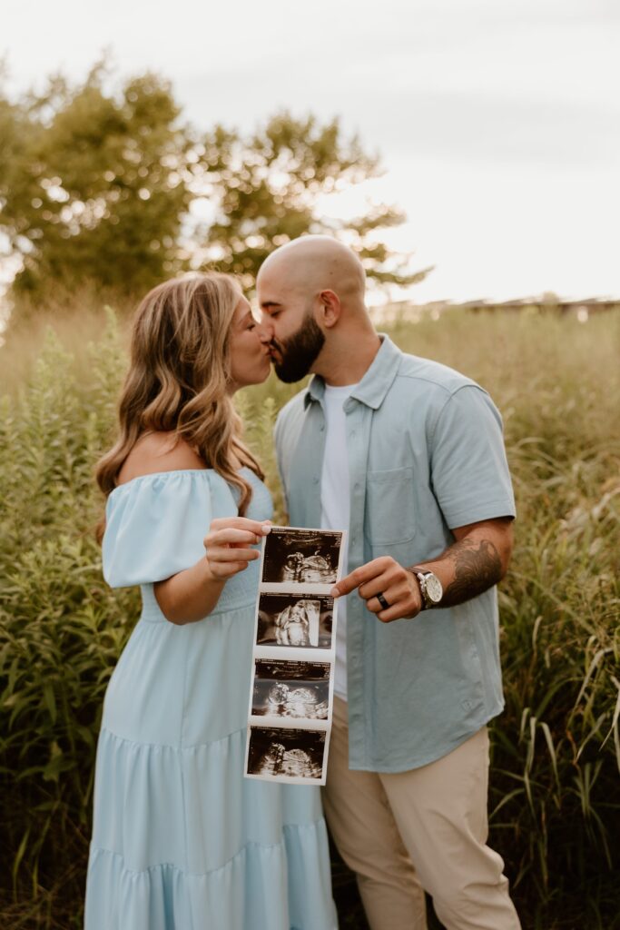 A couple stands in a grassy field, sharing a kiss while holding an ultrasound photo in front of them. The woman wears a light blue, off-the-shoulder dress, and the man wears a matching light blue shirt. The ultrasound photo adds a personal and meaningful touch to this joyful moment as they celebrate their upcoming addition to the family.