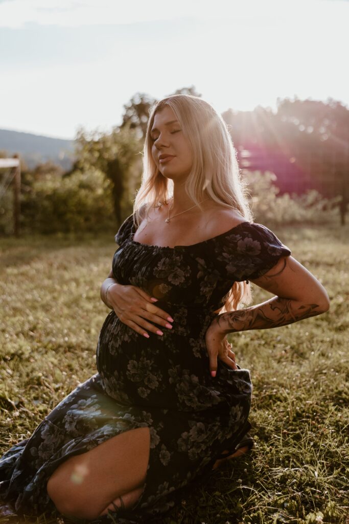 A pregnant woman sits in a sunlit field during golden hour, gently cradling her belly with one hand and supporting her back with the other. She wears a dark floral dress, her long blonde hair glowing in the soft sunlight. The peaceful expression on her face is enhanced by the warm, natural light, creating a serene and radiant maternity portrait.