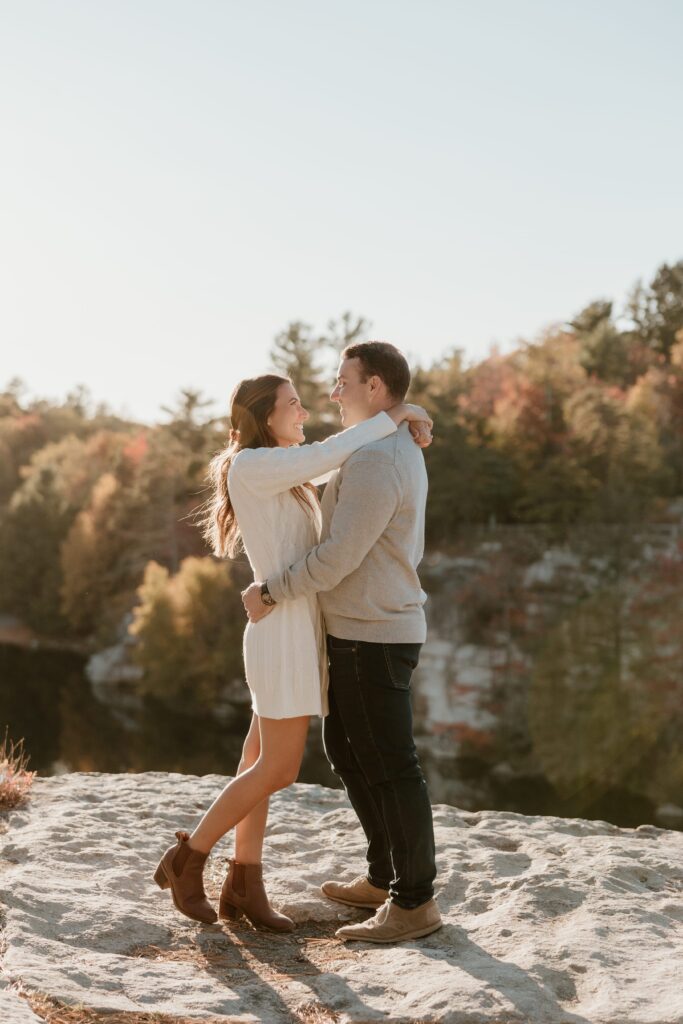 A smiling couple embracing on a rocky overlook during golden hour at Lake Minnewaska, surrounded by autumn foliage and scenic views.