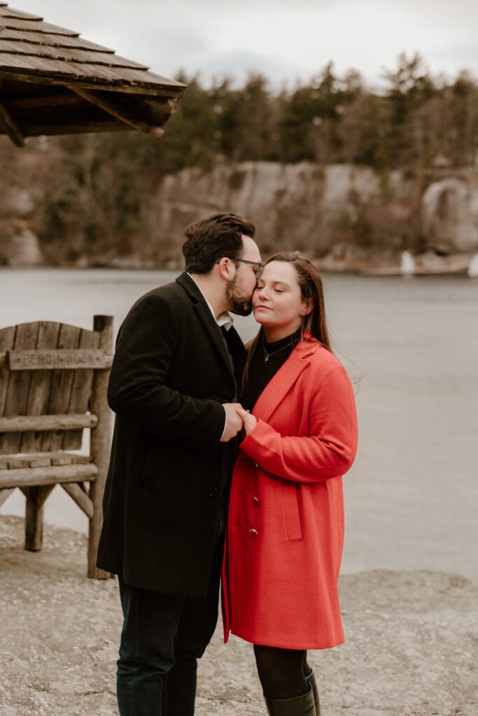 Engaged couple sharing a tender moment by the lake at Mohonk Mountain House, with rustic wooden seating in the background during their winter engagement session.