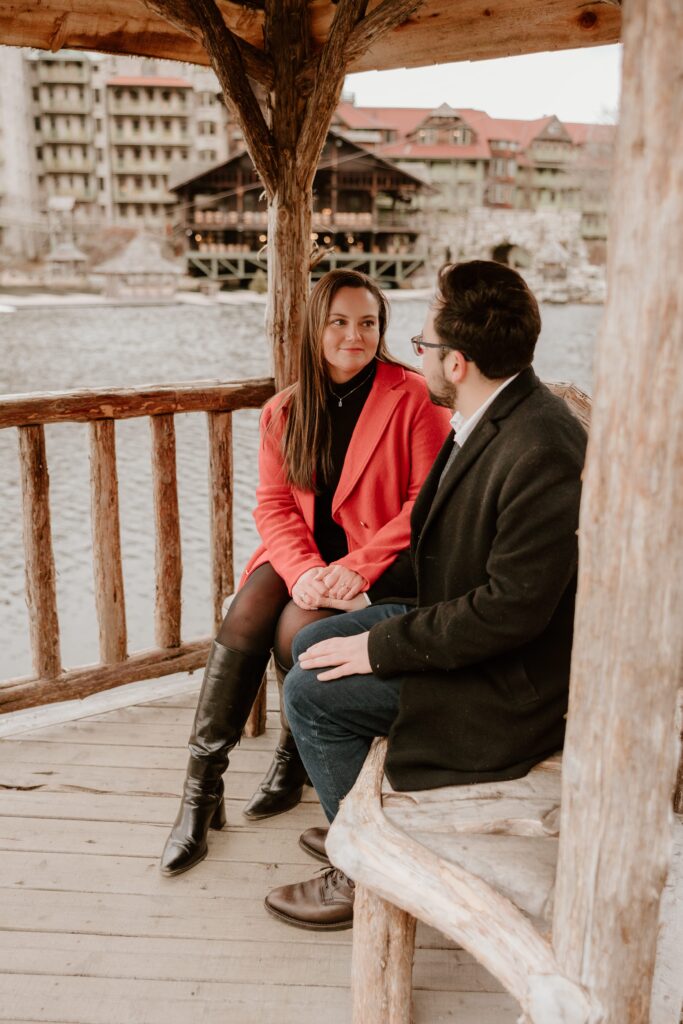 Engaged couple sitting together in a rustic gazebo overlooking the lake, with Mohonk Mountain House in the background during their engagement session.