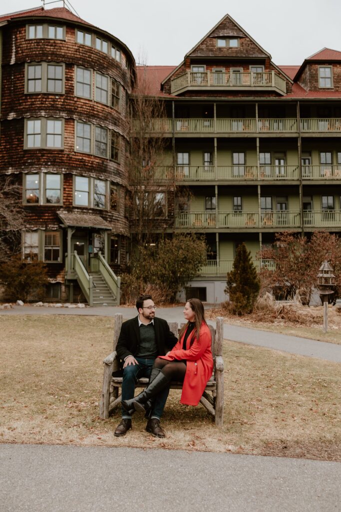 Engaged couple sitting on a wooden bench in front of the historic Mohonk Mountain House building during their engagement session in the Hudson Valley.