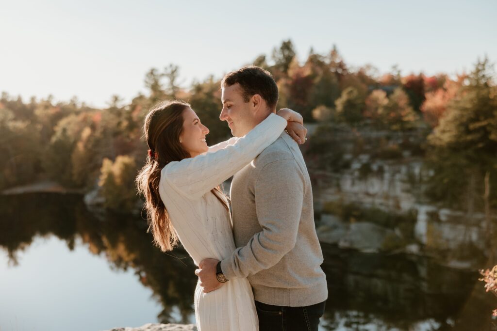 A couple embracing and smiling at each other during their fall engagement session at Lake Minnewaska, surrounded by scenic autumn foliage and reflections.