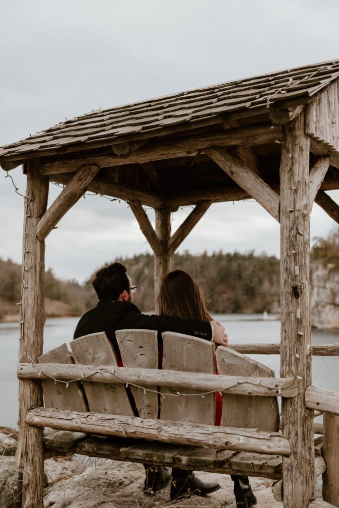 Engaged couple sitting together under a rustic gazebo overlooking the lake at Mohonk Mountain House during their engagement session.