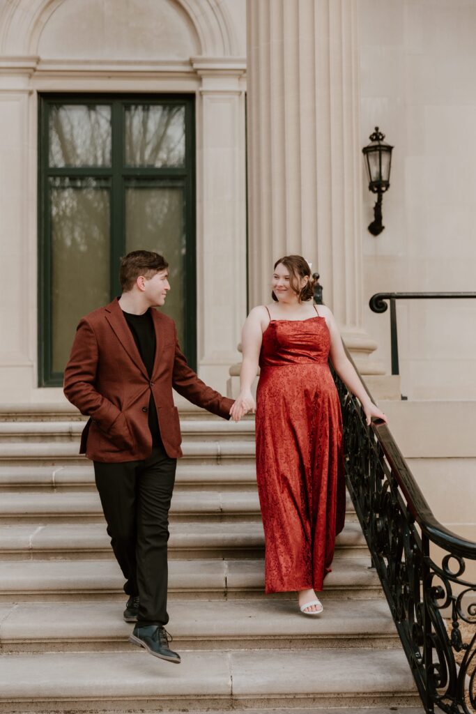 Engaged couple walking down the steps of Vanderbilt Mansion in elegant red and brown attire during their Hudson Valley engagement session.