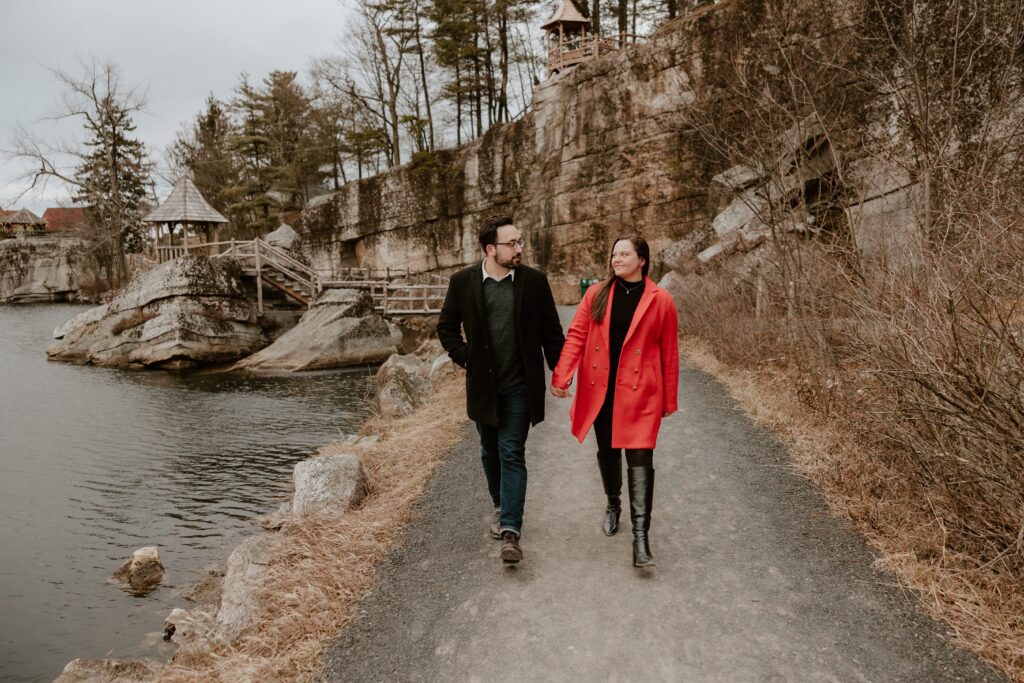 Couple walking hand-in-hand along the lake, surrounded by cliffs and rustic wooden gazebos during their engagement session.