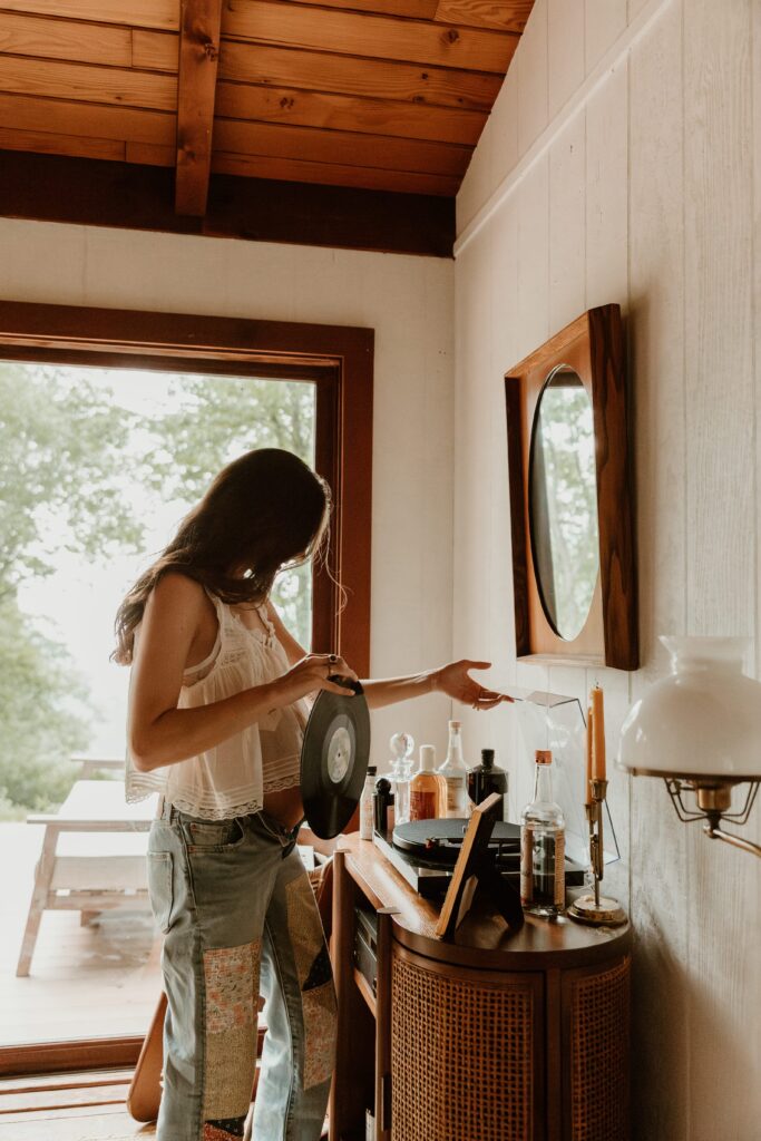 Pregnant woman in a flowy top and patchwork jeans selecting a vinyl record at an Airbnb.