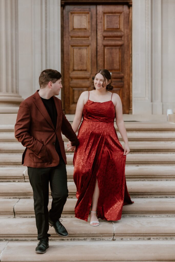 A couple walking hand in hand down the steps of Vanderbilt Mansion in Hyde Park, NY, wearing elegant formal attire during their engagement photoshoot.