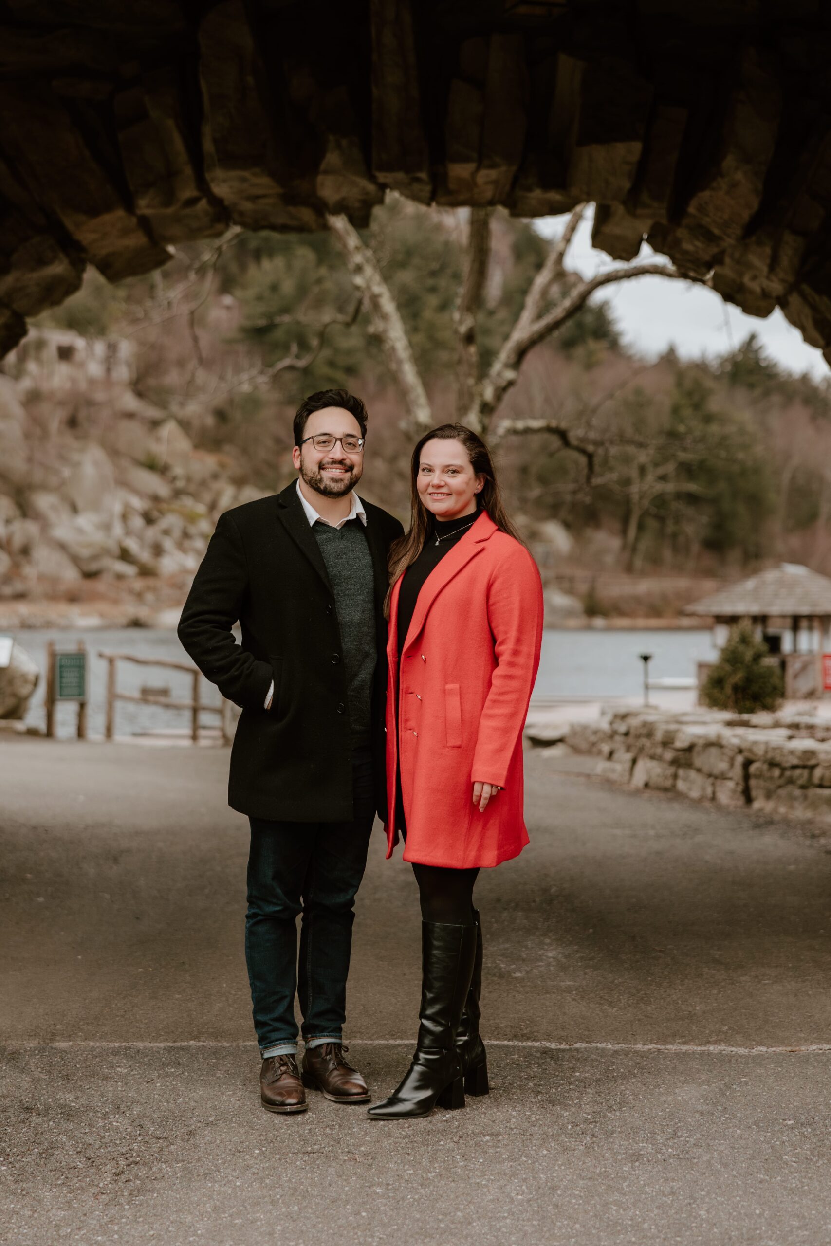 Engaged couple standing under a rustic stone archway at Mohonk Mountain House, smiling and holding hands during a winter engagement session.