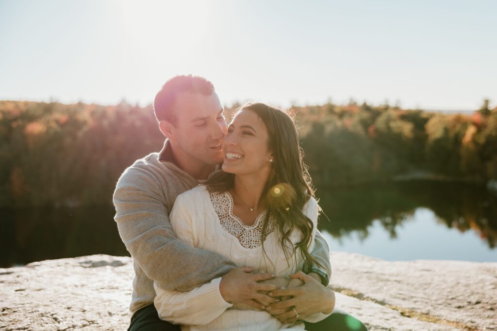 Couple embracing during a golden hour engagement session at Lake Minnewaska, surrounded by fall foliage and reflected in the serene lake.