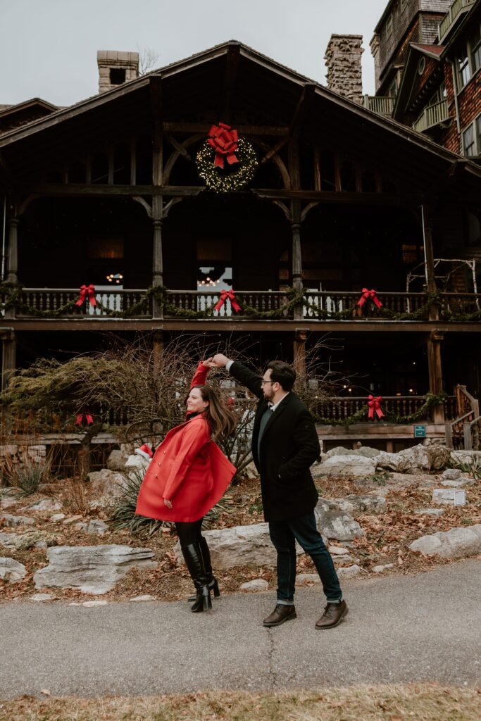 Couple dancing playfully in front of the festively decorated Mohonk Mountain House, featuring wreaths and red holiday bows during their winter engagement session.