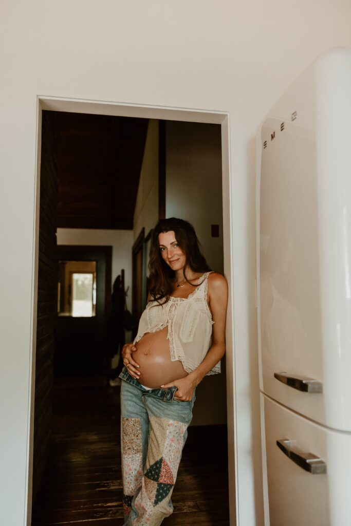 Pregnant woman leaning against a doorway, holding her belly, wearing a bohemian outfit in an Airbnb.