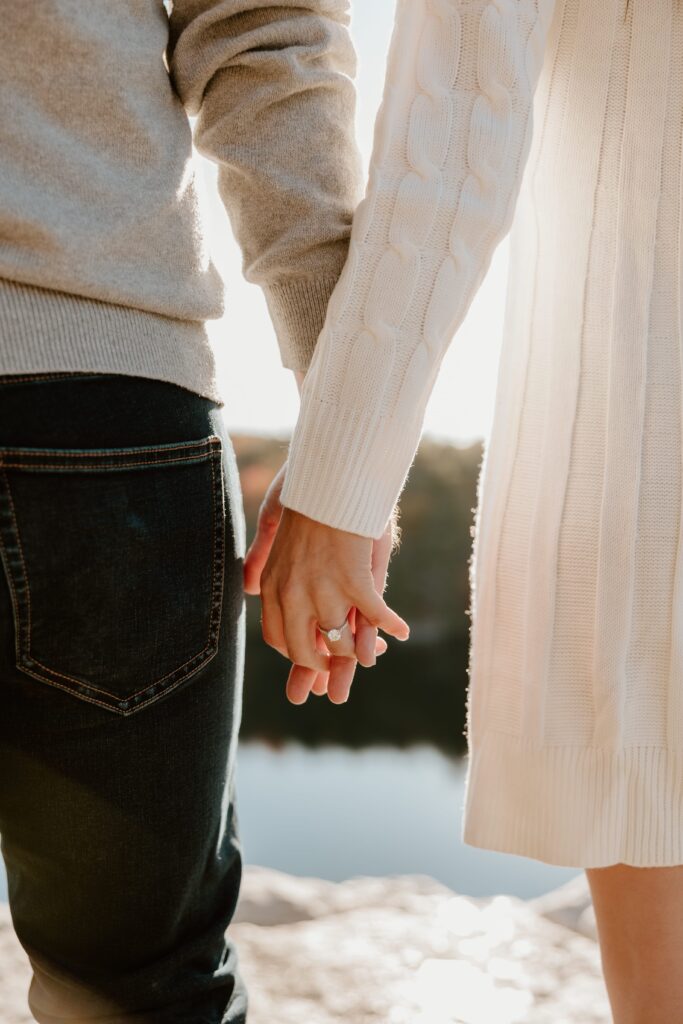 Closeup of a couple holding hands, highlighting a sparkling engagement ring during a golden hour engagement session at Lake Minnewaska.