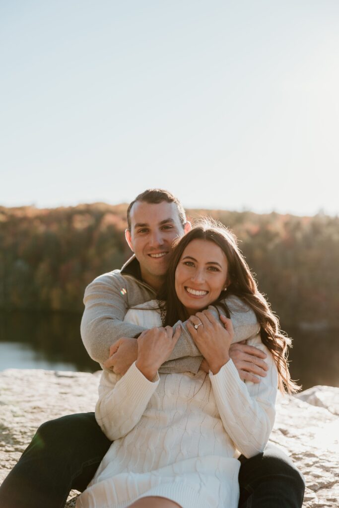 Smiling couple wrapped in a warm embrace during a golden hour engagement session at Lake Minnewaska, with a backdrop of vibrant fall colors.