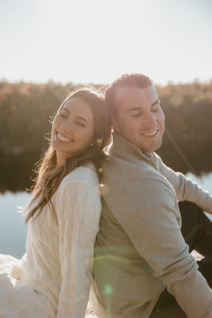 A couple leaning back-to-back, smiling with closed eyes, basking in the golden hour sunlight at Lake Minnewaska during their engagement session.