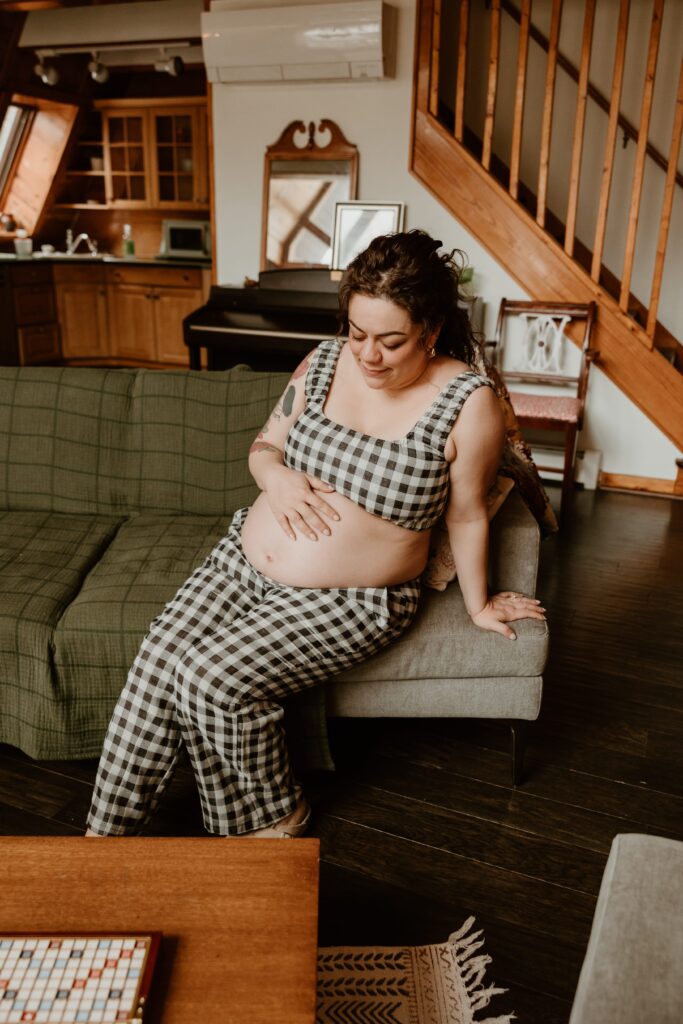 Pregnant mother sitting on a couch in a cozy Airbnb, gently holding her belly and smiling in a checkered outfit.