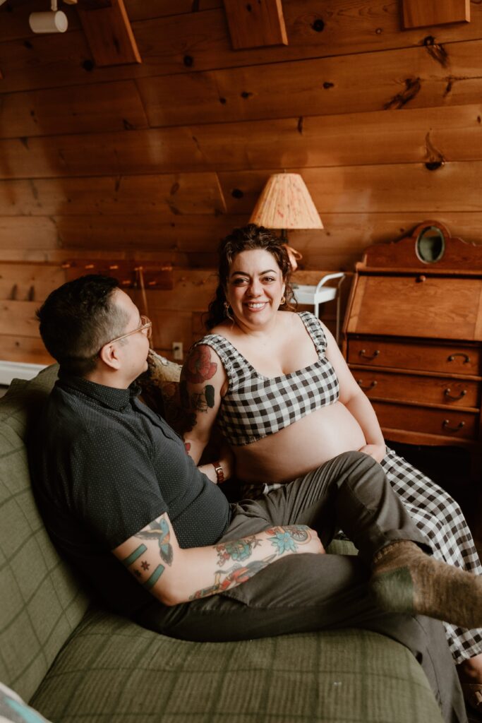 Expecting couple sitting on a green couch in a warm Airbnb setting, smiling and enjoying a heartfelt moment together.