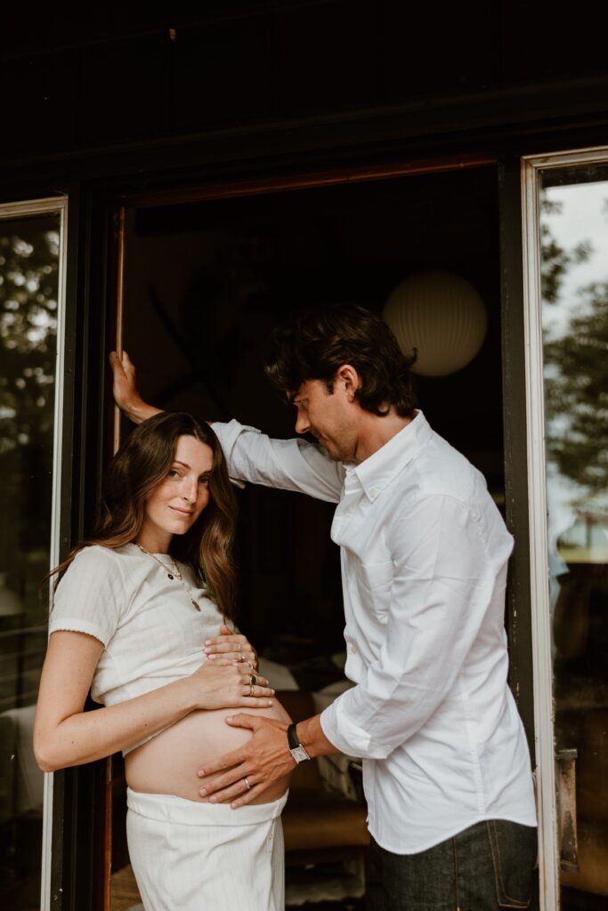 A pregnant couple posing at the front door of a rustic cabin in the Catskills, with the expecting mother leaning against the doorframe and smiling softly at the camera.