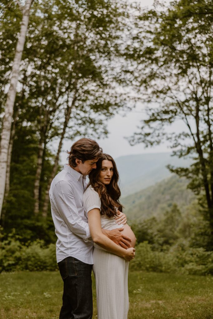 A tender maternity portrait of a couple embracing outdoors, with lush green mountains of the Catskills creating a picturesque backdrop.