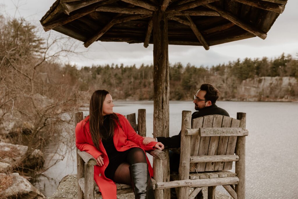 Engaged couple sitting on rustic chairs under the Perch Rock gazebo, holding hands and sharing a moment by the lake at Mohonk Mountain House.