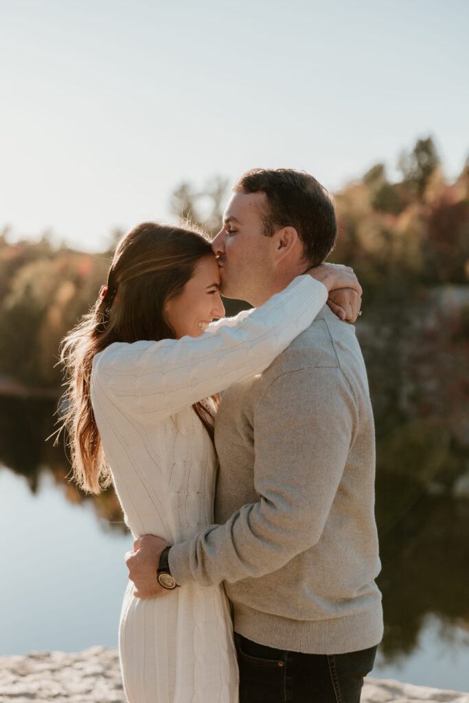 A couple sharing an intimate moment with a kiss on the forehead during golden hour at Lake Minnewaska, surrounded by warm autumn tones.