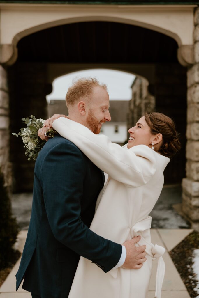 Bride and groom embrace and smile at each other under stone archway at Saint Lawrence O'Toole Catholic Church in Brewster, NY, during their winter ceremony.