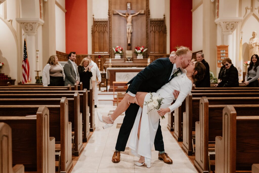 Bride and groom share a dip kiss in the aisle of Saint Lawrence O'Toole Catholic Church in Brewster, NY, with family and friends clapping and smiling in the background.