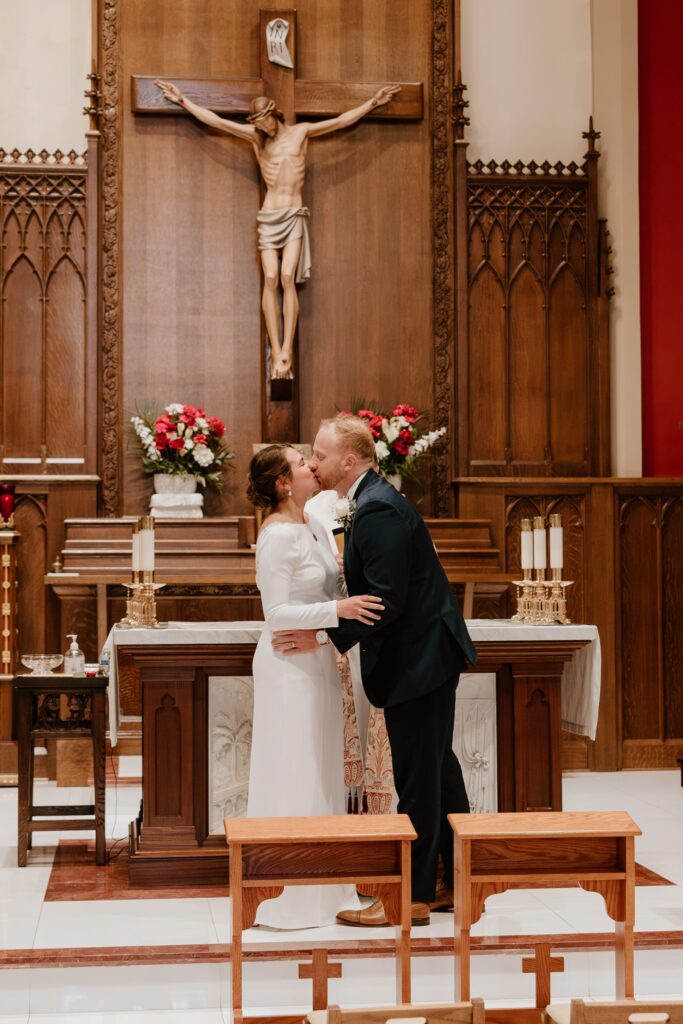 Bride and groom share their first kiss at the altar beneath a large crucifix inside Saint Lawrence O'Toole Catholic Church in Brewster, NY, during their intimate winter ceremony.