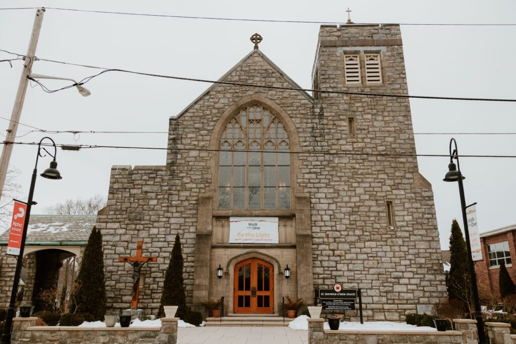 Exterior of Saint Lawrence O'Toole Catholic Church in Brewster, NY, showing stone facade, large arched windows, and wooden doors on a snowy winter day.