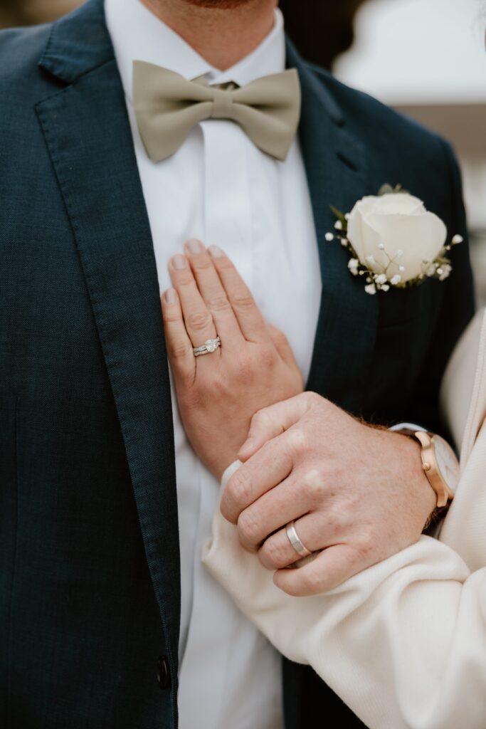 Close-up of bride and groom's hands showing wedding rings, with groom wearing a navy suit, sage green bow tie, and white rose boutonniere during winter church ceremony in Brewster, NY.