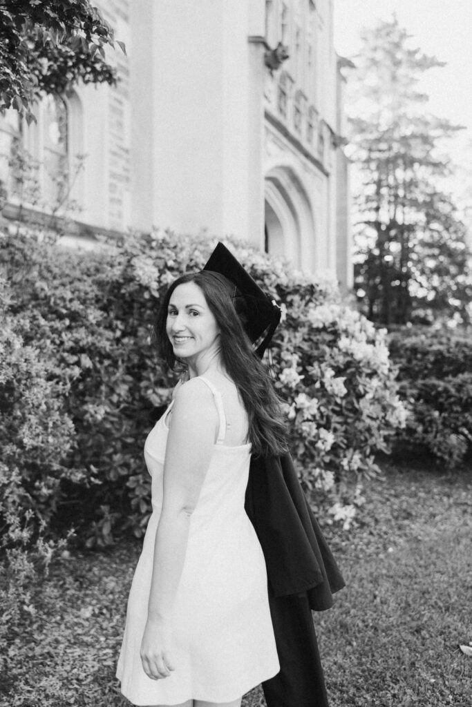 Black and white photo of a Vassar College graduate smiling over her shoulder with her gown draped over her arm, standing in front of campus buildings and blooming bushes.
