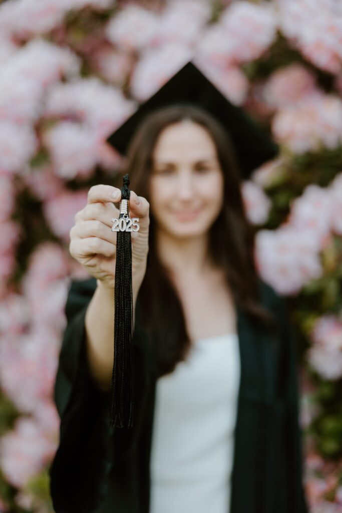 Close-up of a black 2025 graduation tassel held by a student in cap and gown, with pink spring flowers softly blurred in the background at Vassar College.