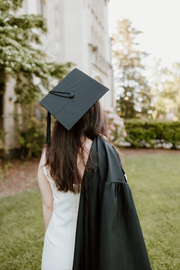 Graduate facing away from the camera with her cap on and gown draped over her shoulder, standing on the lawn at Vassar College during her senior photo session.