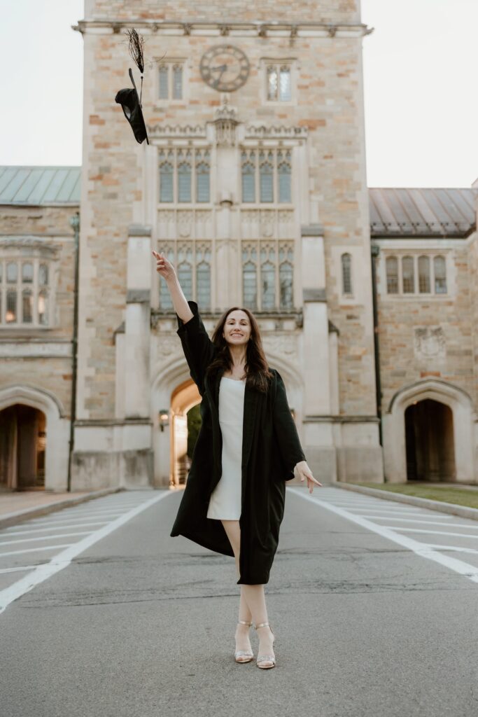 Graduate in cap and gown tossing her cap in front of the iconic Vassar College library building in Poughkeepsie, NY during her senior photo session.