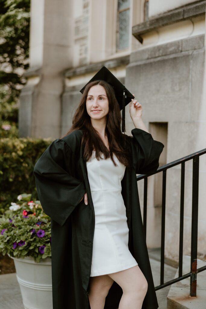 Graduating student in a white dress and black cap and gown posing confidently by the steps of a historic building at Vassar College in Poughkeepsie, NY.