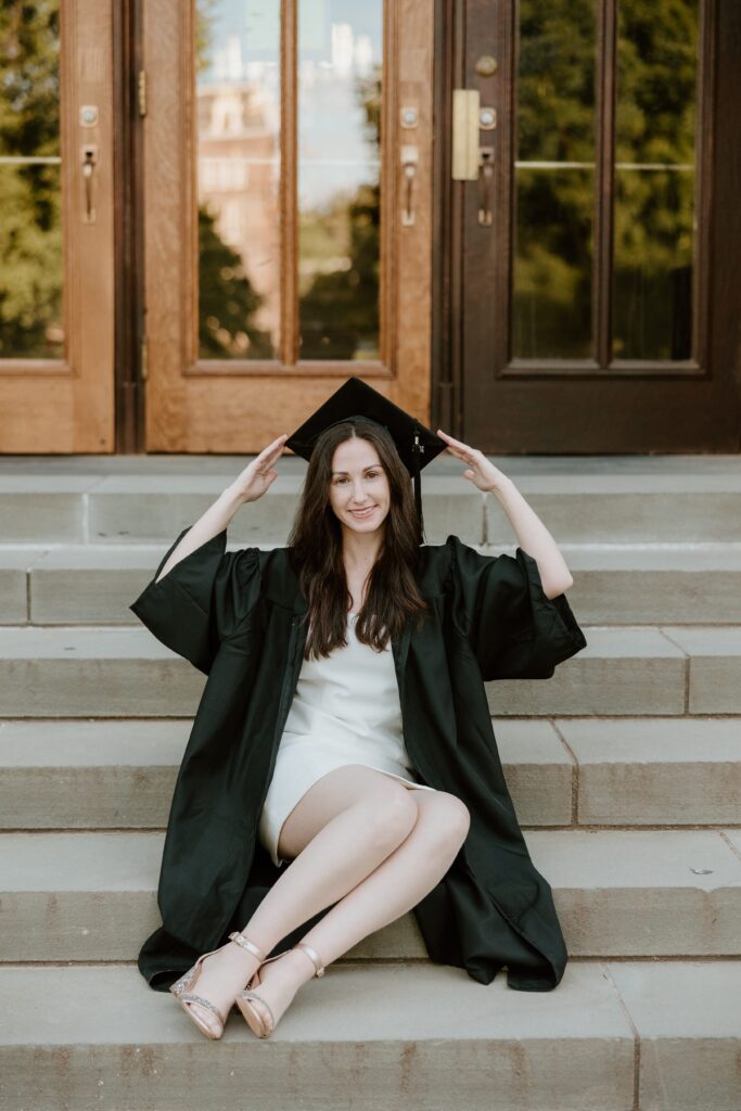 College graduate sitting on the steps outside Vassar College, smiling and adjusting her graduation cap during her senior photo session in Poughkeepsie, NY.