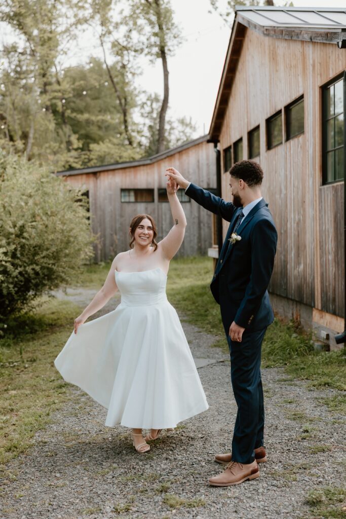 Bride twirls in tea-length wedding dress while groom smiles at her outside rustic barn at Stonehill’s Farmhouse in Accord, NY.