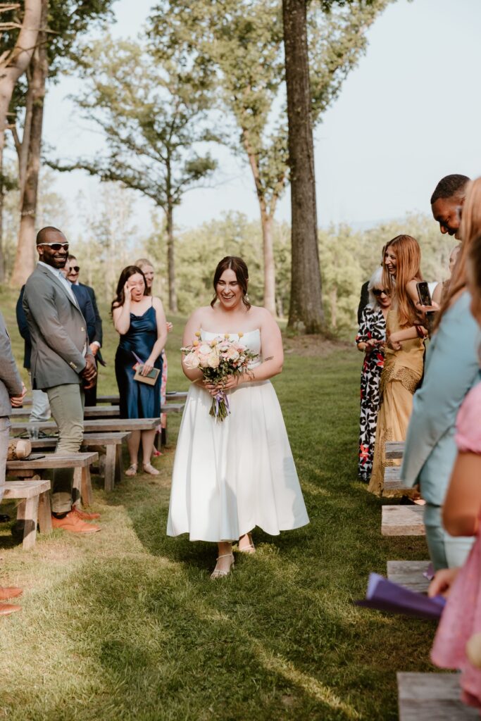 Bride walking down the aisle smiling during outdoor ceremony at Stonehill’s Farmhouse in Accord, NY, surrounded by joyful guests.
