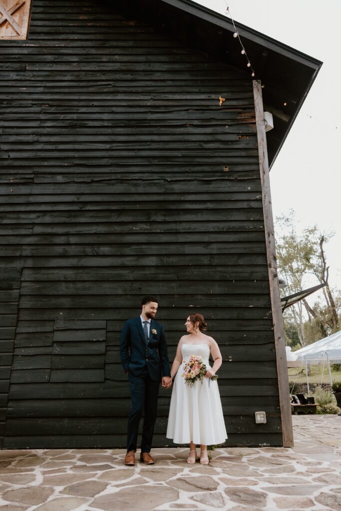 Couple holds hands in front of black barn wall at Stonehill’s Farmhouse wedding in Accord, NY.