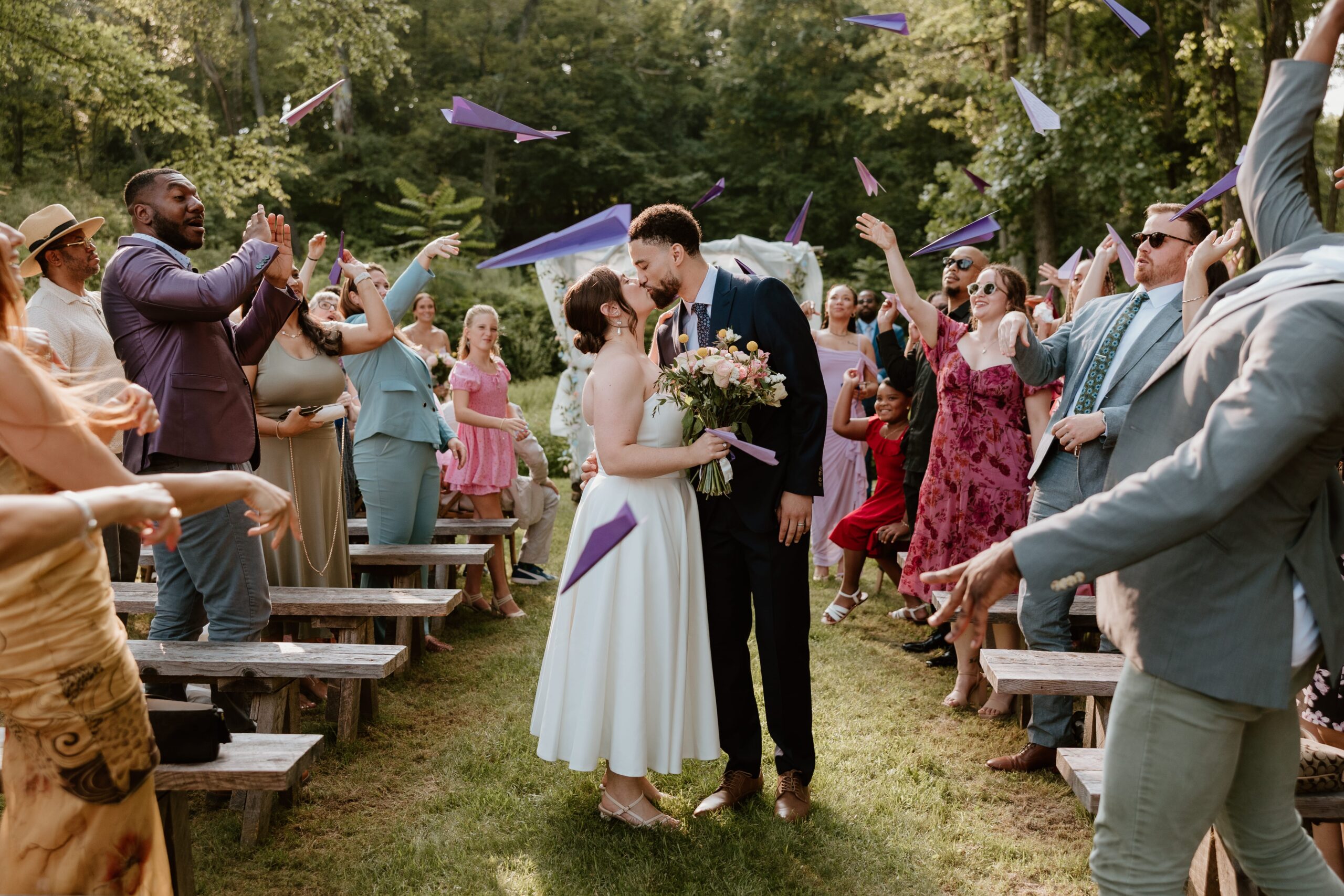 Bride and groom kiss during outdoor wedding ceremony exit while guests throw purple paper airplanes in celebration at Stonehill’s Farmhouse in Accord, NY.