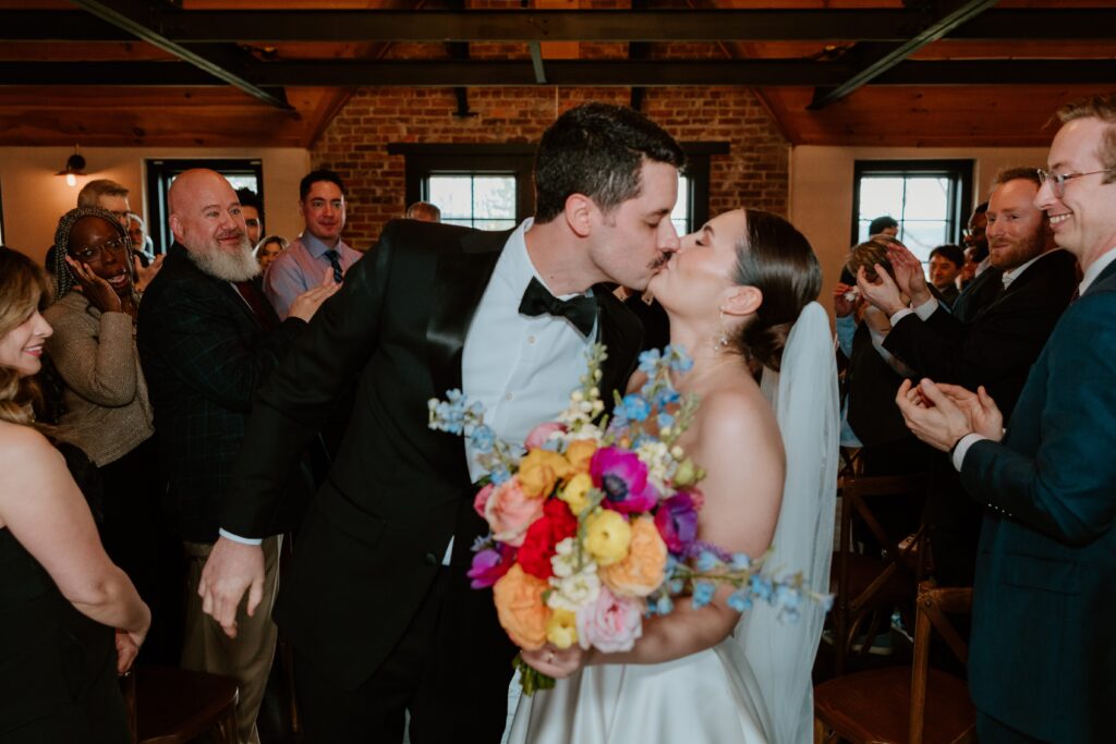 Bride and groom sharing their first kiss during an intimate wedding ceremony at Drowned Lands Brewery in Warwick NY surrounded by guests.